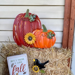 May include: A large red metal pumpkin and a smaller orange metal pumpkin, both featuring green stems and leaves. A yellow sunflower is attached to the pumpkins. A white sign reads: BUT I THINK I LOVE fall MOST OF ALL. A black bird figure sits nearby.