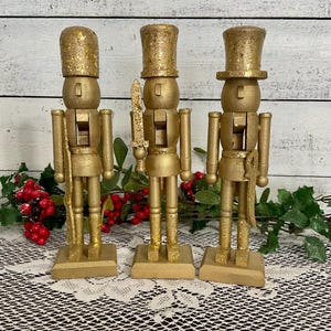 May include: Three gold-painted wooden nutcrackers with glittered hats and bases. Each figure is posed differently, one holding a sword, another a rifle, and the third with hands at its sides. The background includes greenery and a white wooden surface.