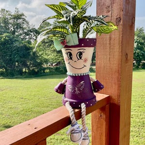 May include: A whimsical, hand-painted terracotta flower pot shaped like a girl, with a purple dress and white polka dots. The pot features a smiling face, a green bow, and a butterfly design. The girl is sitting on a wooden railing, with rope legs and canvas shoes.