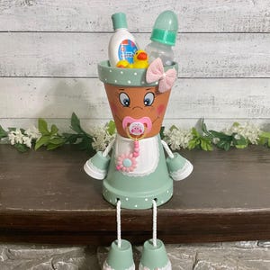 May include: A terracotta flower pot transformed into a baby-themed decorative item. The pot is painted with a face and adorned with a pacifier, a bow, and a green apron. It holds baby essentials like a bottle, lotion, and a rubber duck.