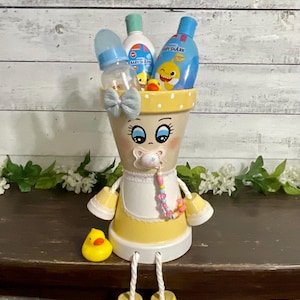 May include: A whimsical, hand-painted baby-themed flower pot. The pot is decorated with a face, pacifier, and bow, and filled with baby products. A rubber ducky sits nearby. The pot is yellow and white.