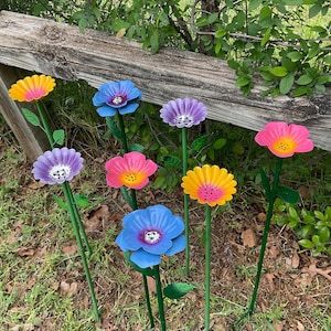 May include: A collection of colorful metal flower garden stakes. The flowers are painted in shades of pink, yellow, blue, and purple with yellow centers. The stakes are green and the flowers are arranged in a garden setting.