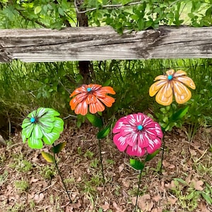 May include: Four colorful metal flower garden stakes. The flowers are in shades of green, orange, yellow, and pink, each with a turquoise center. The stakes are placed in a garden setting with a wooden fence in the background.