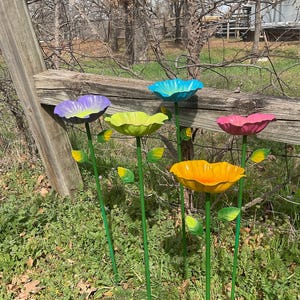 May include: A collection of colorful metal flower garden stakes. Each stake features a different colored flower head, including purple, blue, yellow, pink, and orange. The stakes are green and have attached metal leaves. The flowers are placed in a grassy area next to a wooden fence.