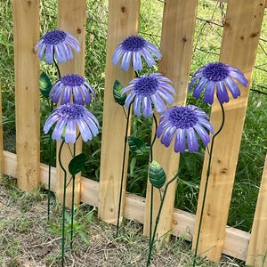 May include: Metal garden art featuring several purple and blue daisy-like flowers with green stems and leaves. The flowers are positioned in front of a wooden fence, creating a decorative outdoor display. The flowers are a vibrant addition to any garden.