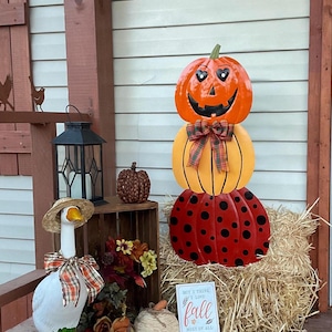 May include: A vertical metal pumpkin stack decoration featuring an orange jack-o'-lantern with heart-shaped eyes, a yellow pumpkin, and a red pumpkin with black polka dots, accented by a plaid ribbon bow and a sign reading But I think I love fall most of all.