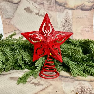 May include: A red metal star Christmas tree topper with a spiral base. The star has cut-out designs and white accents. The topper is placed on a bed of green garland, with vintage paper in the background.