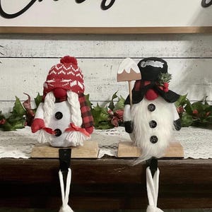 May include: Two Christmas gnome stocking holders. One gnome wears a red and white knit hat and a red and black plaid scarf. The other gnome wears a black hat and holds a wooden shovel. Both have white beards and black button accents.