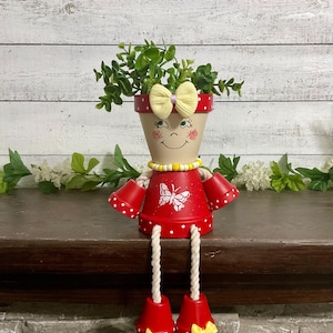May include: A whimsical, handcrafted flower pot doll. The doll features a red body with white polka dots, a cream-colored face with painted features, and a yellow bow. The doll's arms and legs are made of rope, and the pot is filled with greenery.