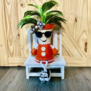 May include: A whimsical terracotta pot transformed into a character, complete with sunglasses, a bow, and a red outfit adorned with daisy accents. The pot is planted with a vibrant green plant and sits on a small white chair, with sneakers dangling.