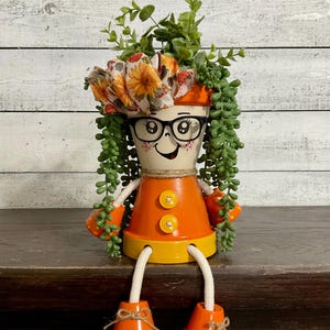 May include: A decorative figurine crafted from stacked terracotta pots painted orange and yellow. The top pot features a hand-painted face with black glasses, while the body has two yellow buttons. It is adorned with a sunflower ribbon and faux greenery.