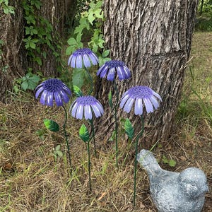 May include: A cluster of purple metal flower garden stakes with green stems and leaves. The flowers have a layered petal design. A gray bird statue is in the foreground, and a tree trunk is in the background.