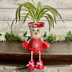 May include: A whimsical, hand-painted terracotta pot shaped like a person, holding a green plant. The red pot features white polka dots, a smiling face, and decorative accents. The arms and legs are also made of terracotta.