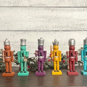 May include: Six wooden nutcracker figurines with different colored paint finishes. The nutcrackers are standing in a row on a wooden surface. The nutcrackers are wearing hats and have painted facial features.