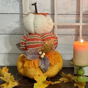May include: A three-tiered decorative pumpkin with a white, orange, and yellow color scheme. The top pumpkin is white, the middle one is orange and brown striped, and the bottom is yellow. Autumn leaves and a lit candle complete the fall theme.