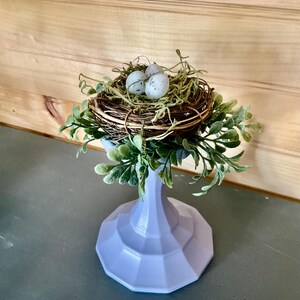 May include: A decorative centerpiece featuring a small bird's nest with three speckled eggs, nestled on a bed of dried twigs and greenery. The nest sits atop a light purple, faceted pedestal, creating a charming spring or Easter theme.