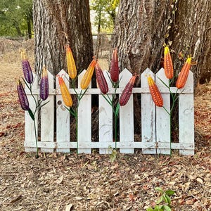 May include: A collection of ten decorative metal corn stalks featuring vibrant, textured kernels in shades of purple, yellow, orange, and red. Each stalk is attached to a thin green metal stem, arranged in a row against a white wooden picket fence.