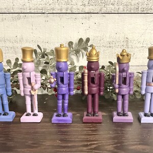 May include: Six wooden nutcracker figurines with gold crowns and hats. The nutcrackers are painted in shades of purple, blue, and lavender. They are standing on a wooden surface.