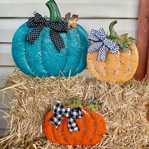 May include: Three metal pumpkin decorations with embossed leaf patterns and glitter accents. The largest is teal with a black and white polka dot bow, the medium is orange with a polka dot bow, and the smallest is bright orange with a buffalo check bow.