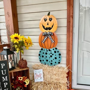 May include: A vertical metal wall hanging featuring three stacked pumpkins. The top pumpkin is orange with heart-shaped eyes and a jack-o'-lantern face. The middle pumpkin is orange with a black and white buffalo check bow. The bottom pumpkin is teal with black polka dots.
