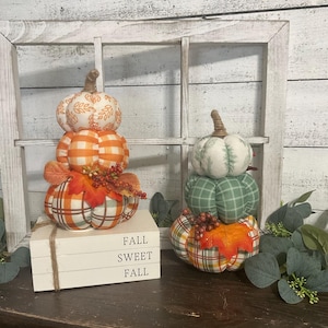 May include: Two stacked fabric pumpkin sets featuring orange, white, and green plaid and leaf patterns, accented with faux berries and leaves. The pumpkins sit atop a stack of three white wooden blocks with the text FALL SWEET FALL.