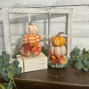 May include: Two stacked fabric pumpkin sets featuring orange, white, and green plaid and leaf patterns. The left set sits on three white books with the text FALL SWEET FALL. Both pumpkin stacks are accented with artificial autumn leaves and berries.