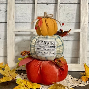 May include: A three-tiered pumpkin decoration with a rustic aesthetic. The top pumpkin is orange, the middle one is plaid, and the bottom one is red. The middle pumpkin has a label that says "PUMPKINS". The decoration is adorned with fall foliage.