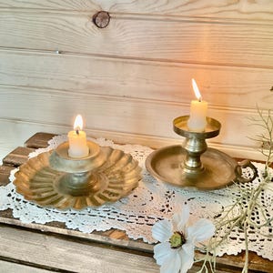 May include: Two antique brass candle holders with lit candles. One has a scalloped base, the other a round base with a handle. A white lace doily and a white flower are also visible.