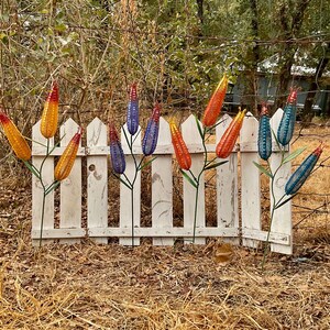 May include: A collection of eleven decorative metal garden stakes shaped like corn cobs, featuring a mesh texture and vibrant colors including yellow, orange, red, purple, and blue. Each stake has a thin green metal stem and a small curled wire top.