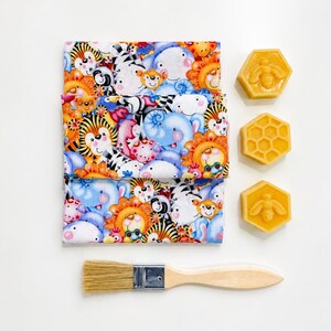 May include: A folded piece of fabric featuring a colorful cartoon animal print with tigers, zebras, and elephants, accompanied by three yellow hexagonal beeswax blocks with bee and honeycomb patterns and a wooden brush with light tan bristles.