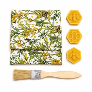 May include: A folded white fabric featuring a pattern of green leaves, yellow flowers, and small bees, accompanied by three hexagonal yellow beeswax blocks and a wooden brush with light-colored bristles and a metal ferrule.