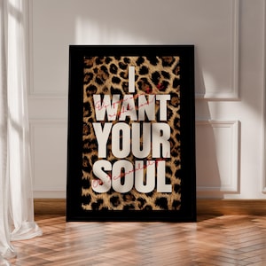 May include: A rectangular framed wall art piece featuring a bold leopard print background. Large, white, block-lettered text reads "I WANT YOUR SOUL" centered on the print. Smaller, thin, red cursive text is layered over the white lettering.