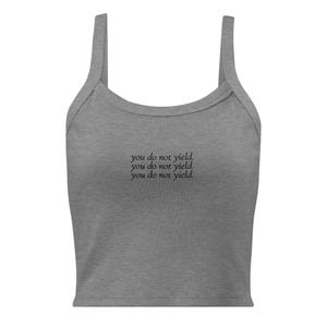 May include: A heather gray sleeveless crop top with a scoop neckline and thick shoulder straps. The center of the chest features the text "you do not yield." printed in a black, serif font, repeated in three stacked lines across the front.
