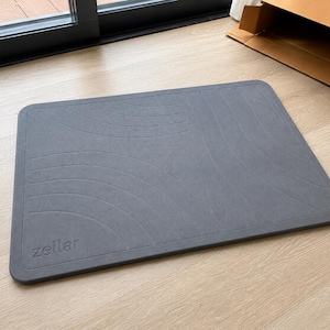 May include: A rectangular dark gray bath mat with rounded corners and a textured surface featuring embossed curved lines. The word zellar is debossed in the bottom left corner of the mat. The mat has a solid, flat, and firm appearance.