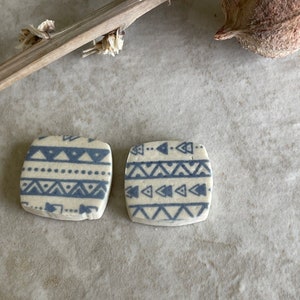 May include: Two square ceramic tiles with rounded corners, featuring a white base color and a repeating pattern of blue horizontal stripes, triangles, and dots. The tiles have a matte finish and are positioned side by side on a textured surface.