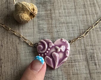 May include: A necklace featuring a glossy, light purple heart-shaped pendant with embossed swirl and butterfly patterns. The pendant is attached to a gold-toned metal chain with elongated oval links. The heart has a smooth, glazed finish.