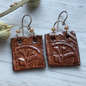 Puede incluir: Un par de pendientes cuadrados marrones con acabado brillante. Cada pendiente presenta un diseño floral en relieve y cuelga de un gancho dorado. Pequeñas cuentas de tonos neutros están unidas a los ganchos.