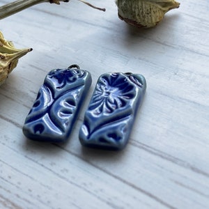 Puede incluir: Dos colgantes rectangulares de cerámica azul con intrincados diseños florales y de volutas. Cada colgante tiene un pequeño agujero para ensartar. Los colgantes se muestran sobre una superficie de madera clara.