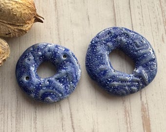 May include: Two blue ceramic donut-shaped beads with a speckled glaze. The beads have a hole in the center and are perfect for jewelry making.