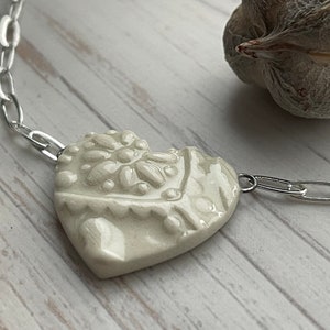 Op de afbeelding: Een crème-kleurige hartvormige hangerketting met een zilveren ketting. De hanger heeft een textuur bloemenontwerp. De ketting wordt gepresenteerd op een licht houten oppervlak, met een gedroogde zaaddoos op de achtergrond.