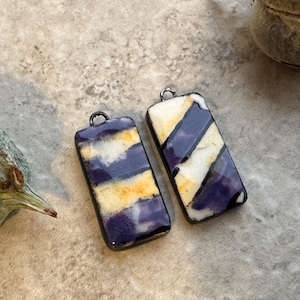May include: Two rectangular pendants featuring a smooth, glossy finish with diagonal stripes of deep purple, cream, and golden yellow. Each pendant is encased in a dark metal frame and includes a small circular loop at the top for attachment.