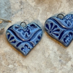 May include: Two blue ceramic heart pendants with floral patterns and a metal loop for hanging. The hearts have a glossy finish and are in a close-up shot. The pendants are likely for jewelry making or crafting.
