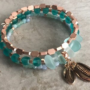 May include: A multi-strand wrap bracelet featuring rose gold-toned hexagonal beads, frosted teal and light blue glass beads, and two dangling metallic rose gold-toned cowrie shell charms. The design incorporates a spiral structure with alternating bead textures and colours.