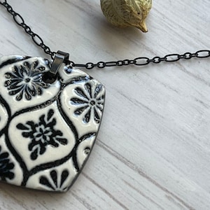 May include: A black chain necklace with a square pendant. The pendant is white with a black floral pattern. The necklace is on a light-colored wooden surface.