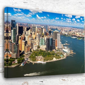 May include: A rectangular canvas wall art print featuring an aerial view of the New York City skyline, including skyscrapers, Battery Park, and the surrounding harbor waters under a bright blue sky with scattered white clouds on a sunny day.
