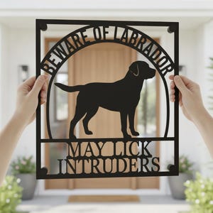 May include: A black metal rectangular sign featuring a silhouette of a Labrador dog in the center. The text "BEWARE OF LABRADOR" is arched above the dog, and the text "MAY LICK INTRUDERS" is positioned in two lines below the dog silhouette.