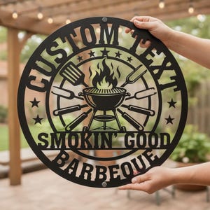 Könnte beinhalten: Ein rundes schwarzes Metallschild mit einer zentralen Grillgrafik samt Flammen, umgeben von Kochutensilien. Der Text lautet CUSTOM TEXT oben, SMOKIN' GOOD in der Mitte und BARBEQUE unten, verziert mit Sternen.