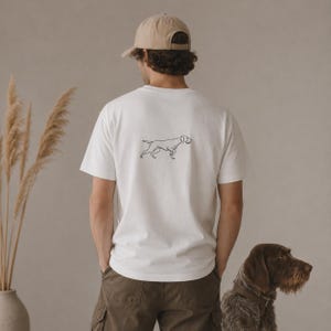 Peut inclure: Un t-shirt blanc à manches courtes avec une illustration au trait noir représentant un chien dans le dos, porté avec une casquette beige et un pantalon cargo vert olive. Le t-shirt est porté à côté d'un chien brun et d'herbes séchées.