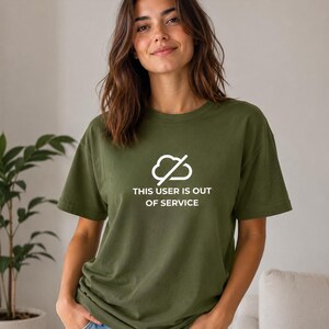 Op de afbeelding: Een olijfgroen T-shirt met korte mouwen en een witte afbeelding van een wolk met een diagonale streep erdoorheen. Onder de afbeelding staat de tekst "THIS USER IS OUT OF SERVICE" in een strak, wit, schreefloos lettertype.