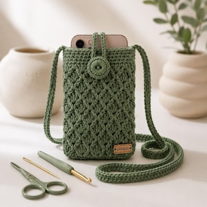 Può includere: Una custodia a tracolla per telefono lavorata all'uncinetto in verde salvia con un motivo a punto conchiglia testurizzato, chiusura con bottone abbinato e tracolla lunga. Una piccola etichetta rettangolare in legno con la scritta "HANDMADE" è fissata sul davanti, accanto a un uncinetto e un paio di forbici.
